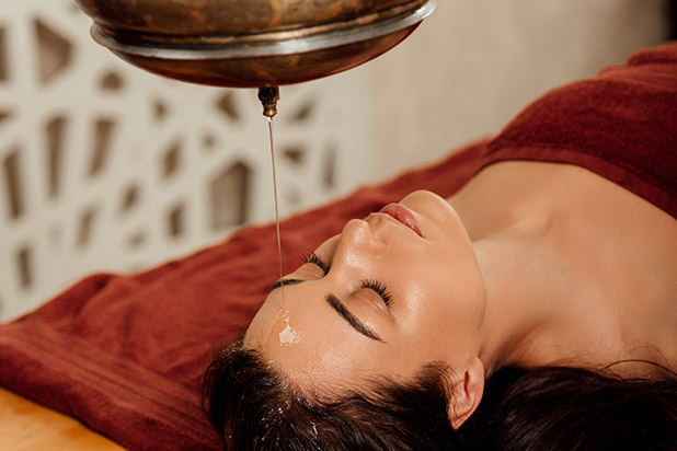 relaxed young woman lying under shirodhara vessel during ayurvedic procedure